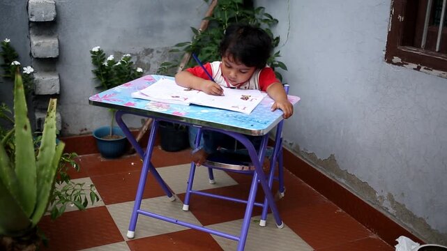 Elementary age cute child of Indian ethnicity sitting on chair near brick wall at home and studying book and writing alphabets in notebook video portrait close up.