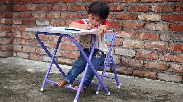 Elementary age cute child of Indian ethnicity sitting on chair near brick wall at home and studying book and writing alphabets in notebook video portrait close up.