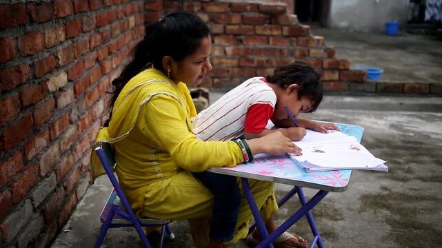 Elementary age cute child and his mother of Indian ethnicity sitting on chair near brick wall at home and studying book and writing alphabets in notebook video portrait together close up.