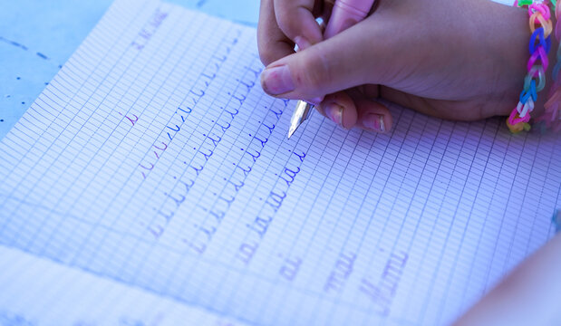 Close-up of a child practicing cursive handwriting with a classic fountain pen and blue ink in a squared notebook, learning to write letters at primary school.