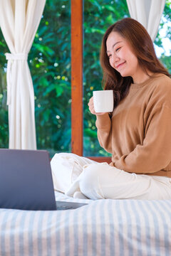 Vertical portrait image of a woman working on laptop computer and drinking coffee while sitting on a bed at home