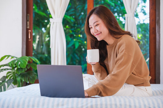 Portrait image of a woman working on laptop computer and drinking coffee while sitting on a bed at home