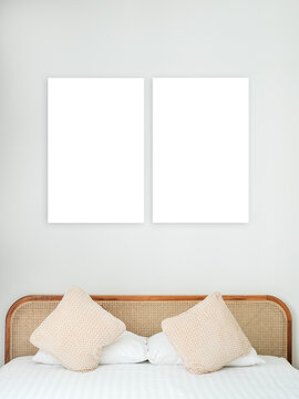 Two blank vertical white frames on soft gray wall above bed with pillows and woven rattan headboard, ideal for poster mockups, wall art presentation, photography and interior design branding concepts.