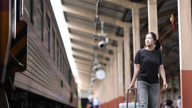 Asian woman with luggage walking on a train station platform. Young woman pulling a rolling suitcase while walking along a platform at a railway station. Concept of travel, vacation, and tourism.