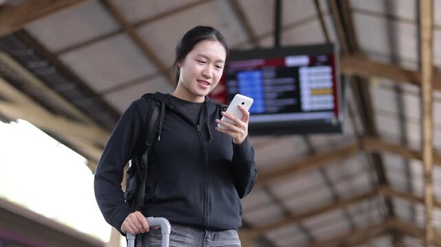 Asian traveler using her smartphone at the train station. Young Asian woman with luggage and a backpack checking her smartphone. A happy female tourist smiling while waiting at a transport hub.