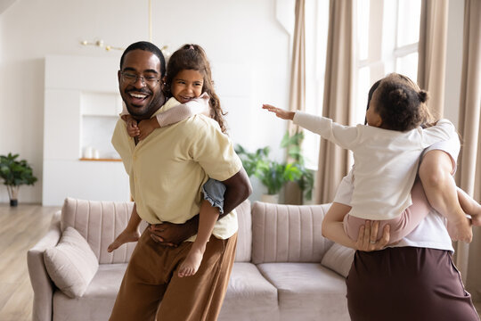 Cheerful mixed-race couple and their two little biracial children having fun at home, running in living room, giving piggyback rides, playing active games together on weekend. Family time and love
