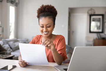 Fototapeta premium Cheerful African woman reading printed document seated at desk with laptop and smartphone, smiling, enjoying professional or personal achievement, bank loan acceptance, reward or great business news