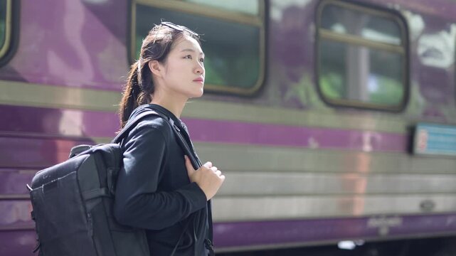 Young Asian woman with backpack waiting for a train. Young Asian woman traveler with a backpack looking around while waiting for the train at the station. Concept of travel and adventure.