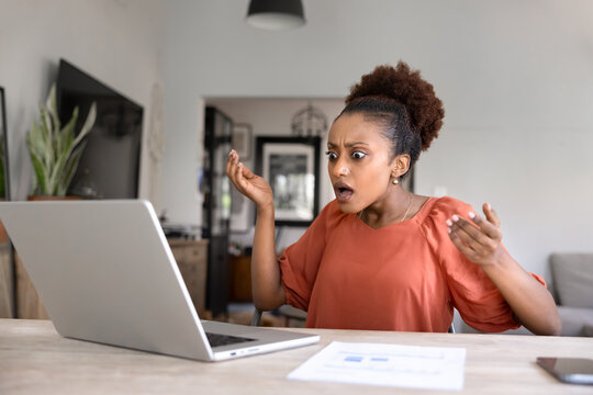 African woman staring at laptop screen with shocked expression, reacts to surprising error with hands raised and open mouth, troubled due to device malfunction or bug, lost information, tech failure