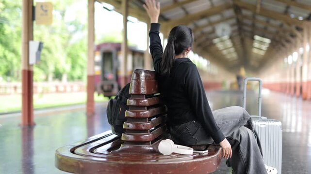 Young woman waving goodbye at a train station platform. Happy young Asian woman sitting on a wooden bench at a train station platform, waving her hand to say goodbye to someone on the train.