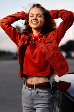 Young woman in a red jacket smiling outdoors. She is enjoying a sunny day near a car. Portrait of a happy stylish female model with closed eyes.