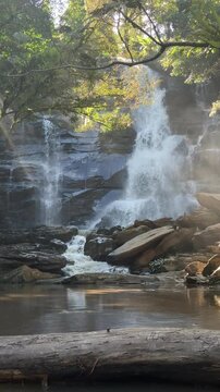 Vertical slow motion footage of a powerful waterfall cascading over dark rocks in a lush forest, with splashing water, mist and reflections creating a dramatic natural scene.