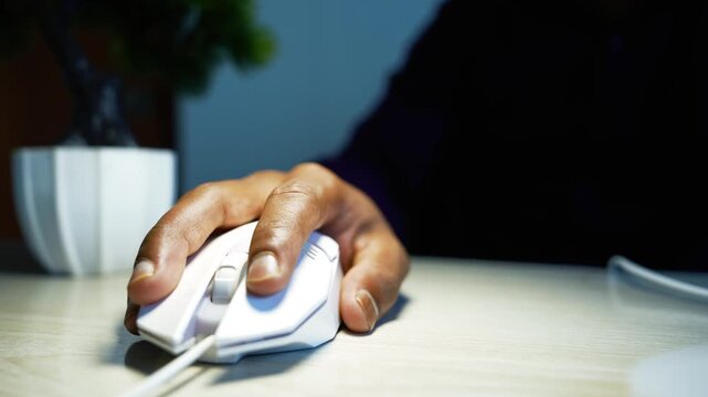 Hand using a computer mouse at an office desk, Close-up of a hand using a white computer mouse on a light desk, with a dark office background and soft natural light