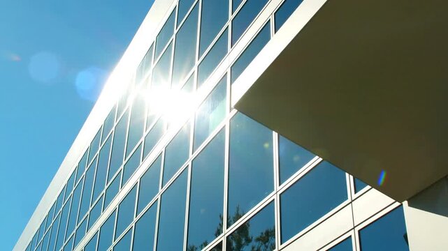 Low angle view of a modern commercial office building facade with blue glass window panes reflecting the clear sky and green trees under bright sunlight during the daytime.
