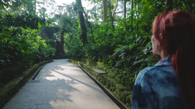 Young woman with pink hair walking along a paved path in a lush green tropical forest