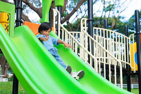 Active young Asian boy having fun sliding down a bright green slide at an outdoor playground. Healthy childhood and weekend recreation concept.