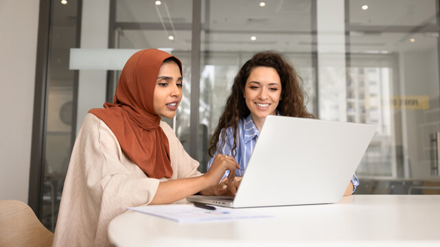 Two multicultural businesswomen collaborating on laptop, reviewing project data, planning strategy during business meeting in corporate office. Communication, teamwork, and professional partnership