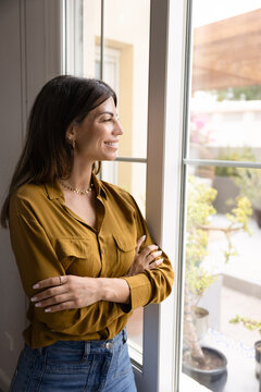 Pensive adult Hispanic woman standing with arms-crossed next to window, smiling, looking outside, spending carefree weekend alone at home, reflecting, thinking, enjoying leisure and personal wellbeing