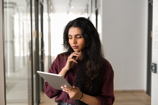 Thoughtful Indian businesswoman working online using digital tablet stand in office corridor, thinking on business information, concentrating on corporate tasks management. Workflow, modern technology