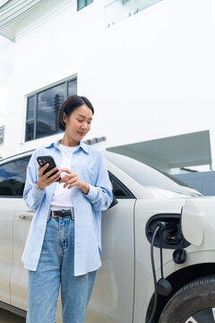 Asian female using a smartphone app while charging her electric car at home. Smart EV home charging, green energy, and sustainable lifestyle. Vertical Shot