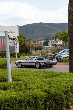 Silver DMC DeLorean Sports Car Rear Three Quarter View Driving Outdoors Near Modern Buildings And Mountain Landscape