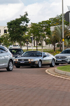 Silver DMC DeLorean Sports Car Front Three Quarter View Driving On Urban Street In City Traffic Near Green Trees
