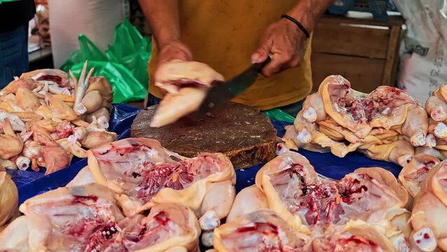A local butcher using a sharp cleaver to cut and prepare fresh raw chicken pieces on a rustic wooden board at an outdoor wet market stall. 4k