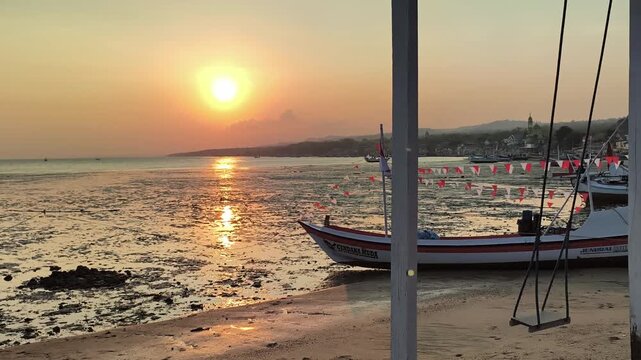 Traditional Indonesian fishing boat and empty beach swing resting on shore at golden hour sunset. 4K Video
