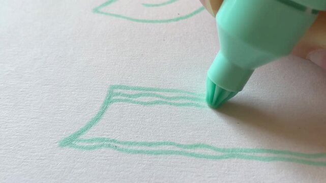 Close-up hand moves mint green felt-tip marker across textured white paper, extending simple outlined drawing with parallel strokes under soft light and shallow focus