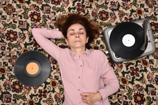 Nostalgic senior woman with closed eyes lying on a Persian carpet enjoying listening to music on a vintage record player. Top view. Concept of people, vintage objects and nostalgic moments.