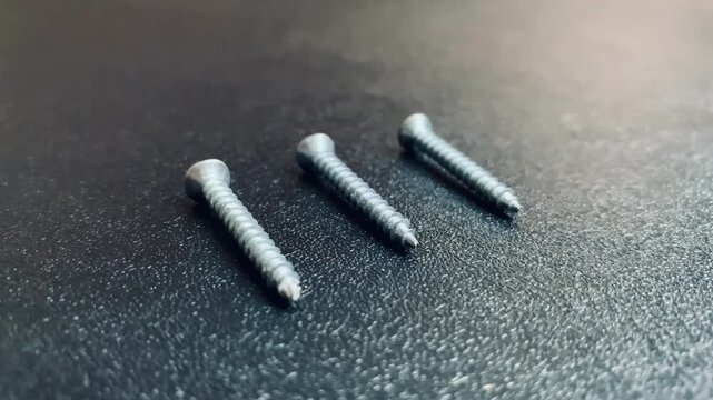 Slow camera pan from left to right showing three metal steel screws arranged in a row on a dark textured background. High angle macro close up 4K video for hardware, home repair, and carpentry.