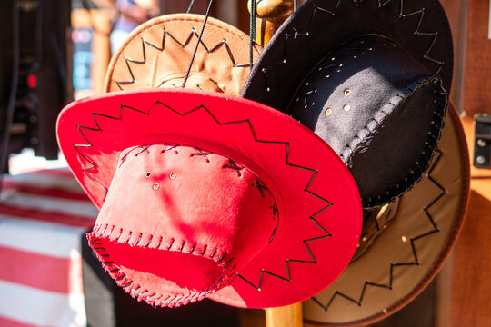 Collection of suede cowboy hats in red, black, and brown with decorative zig-zag stitching on the brim and crown, hanging on a display stand in bright daylight.   