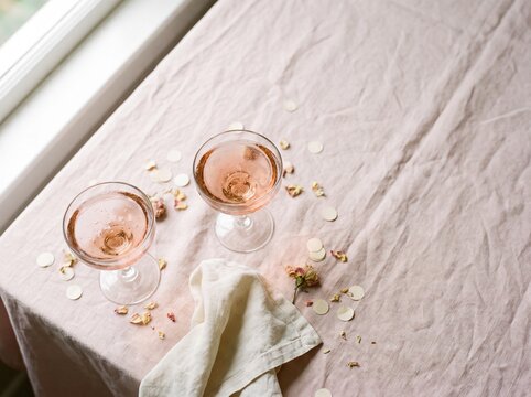 Two coupe glasses of ros&eacute; sparkling drink on pink linen with confetti and petals