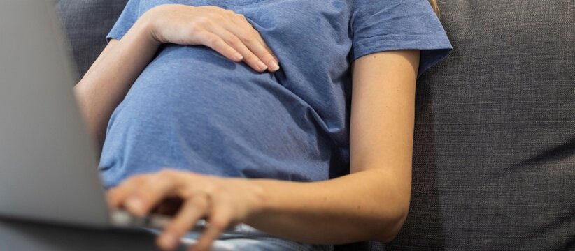 Pregnant woman in blue t-shirt working on laptop on sofa with hand on belly, maternity, remote work and freelance concept