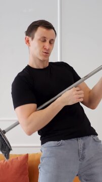 Cheerful Man Performs Rock Concert Playing Floor Mop Like Guitar Inside Modern Living Room. Energetic Dancer Expresses Fun Emotion Through Creative Housekeeping Moves Beside Comfortable Sofa.