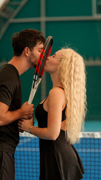 A romantic couple sharing a kiss behind a tennis racket on an indoor court during a match break