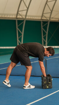 A male tennis player bending over to collect yellow balls from a basket on an indoor court surface