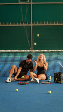 Young couple sitting on a blue indoor tennis court while juggling yellow tennis balls together