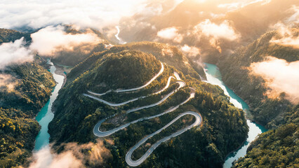 Fototapeta premium An aerial view of a winding mountain road cutting through forested hills beside turquoise rivers, with clouds drifting in valleys at sunrise.