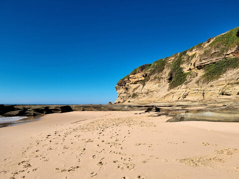 Footprints cross the golden sand of Pebbly Beach towards the layered cliffs and exposed rock platform at Soldiers Point at low tide, Central Coast, New South Wales, Australia.