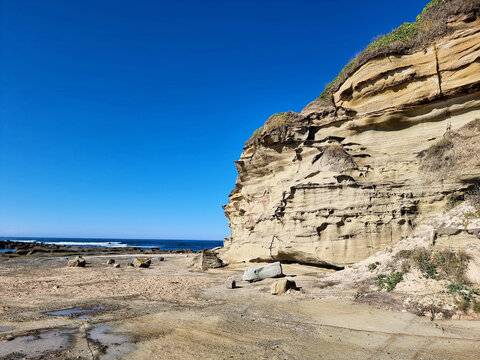 Sunlit coastal cliff with exposed rock layers and sculpted erosion patterns above the rock platform at Soldiers Point at low tide, Central Coast, New South Wales, Australia.