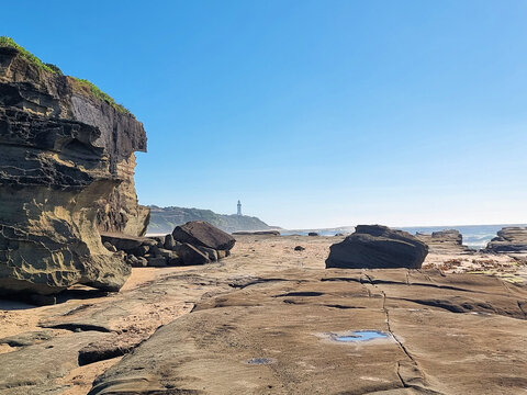 Layered coastal cliffs and scattered boulders frame the rock platform at Soldiers Point, with Norah Head Lighthouse in the distance beneath a clear blue sky, Central Coast, New South Wales, Australia.