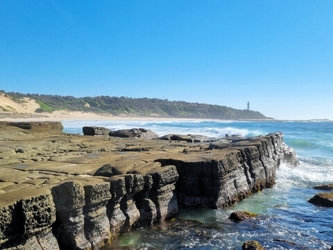 Weathered rock platform and turquoise surf at Soldiers Point, looking towards Norah Head Lighthouse beneath a clear blue sky on the Central Coast of New South Wales, Australia.