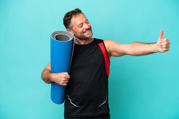 Middle age sport man going to yoga classes while holding a mat isolated on blue background giving a thumbs up gesture © luismolinero