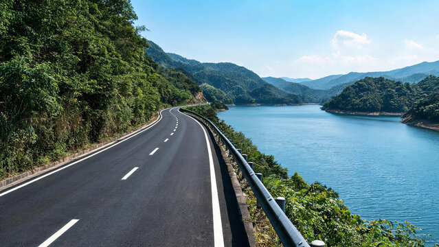 A winding asphalt road runs along a blue lake, surrounded by lush green mountains and forests under a clear blue sky.
