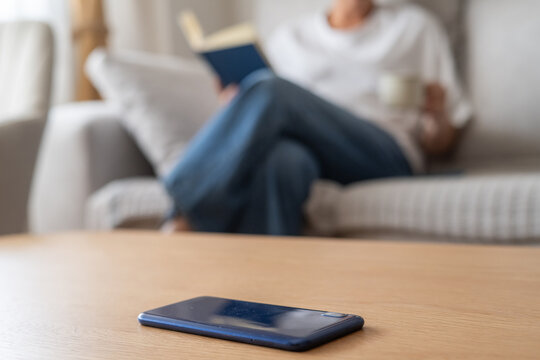 Woman relaxing on a couch reading a book and sipping coffee, intentionally unplugged with her smartphone left on the table for a peaceful digital detox moment at home
