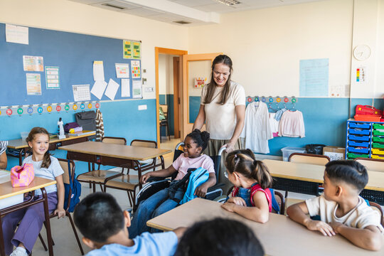 Teacher and diverse students in a classroom, promoting inclusion and support for a smiling girl in a wheelchair