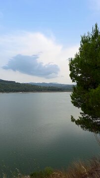 Beautiful scenic outlook of freshwater Beniarres reservoir with turquoise water, pine tree branch in foreground and mountain range in background, Costa Blanca
