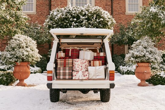 White vintage boxy SUV with open tailgate filled with wrapped gifts and plaid blankets in snowy brick courtyard, festive winter holiday vehicle scene