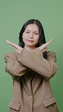 An Asian businesswoman in a beige blazer standing against a green screen and making a stop gesture with crossed arms.